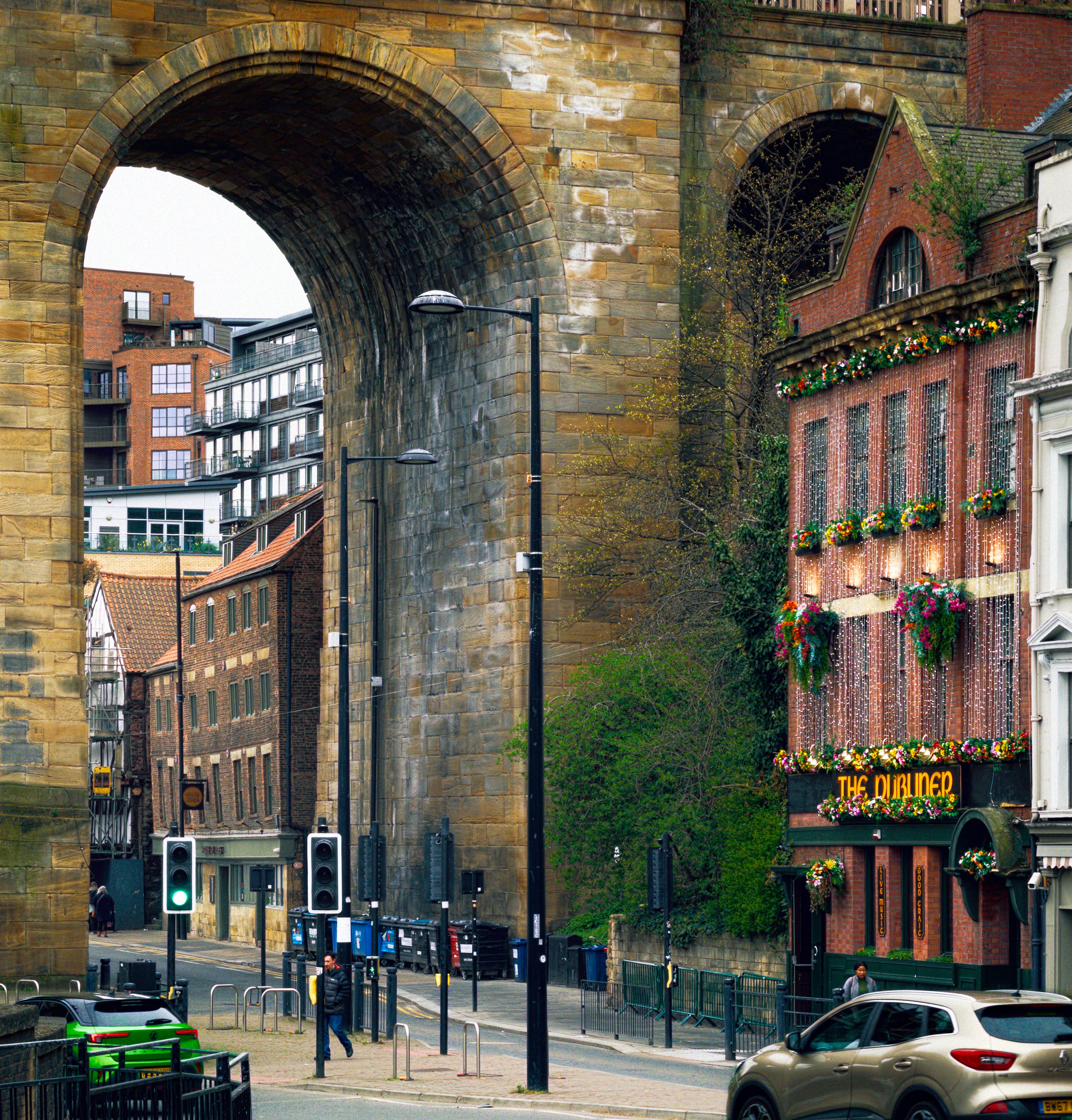 A street scene in Newcastle's Quayside featuring the massive stone arches of a high-level railway viaduct towering over the roadway on the left. Through the largest arch, modern brick apartment buildings with balconies can be seen in the distance against a pale sky. To the right stands a traditional, multi-storey red brick pub named "The Dubliner", its facade brightly decorated with colourful hanging flower baskets and cascading strings of fairy lights above a dark green ground floor. Below, a road with pedestrian crossings, green traffic lights, and tall black lampposts runs beneath the viaduct. A few pedestrians stroll along the pavements, while a bright green car on the lower left and a beige SUV on the lower right navigate the street near a row of commercial wheelie bins nestled against the viaduct's imposing stone pillar.